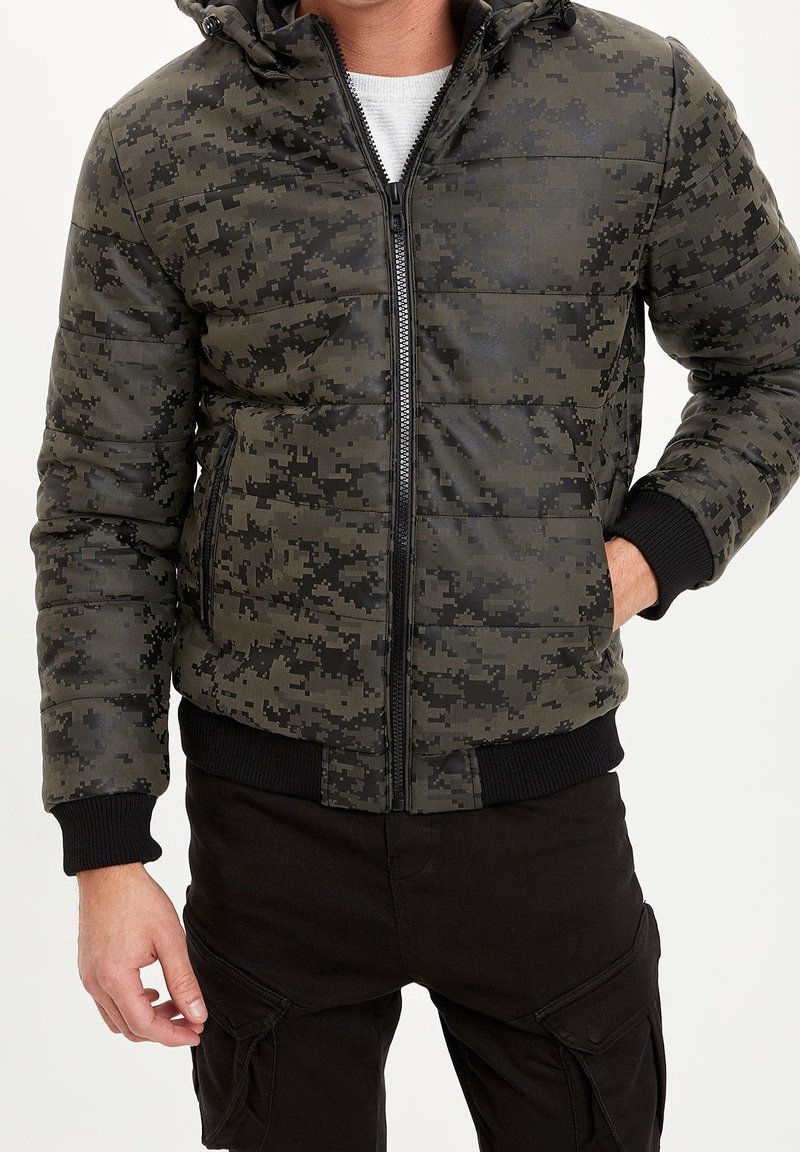 Homme portant une doudoune camouflage numérique verte et noire avec zip devant et poignets et ourlet côtelés noirs, associée à un pantalon cargo noir.