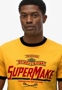 Yellow cotton t-shirt with black ribbed collar, featuring bold red text "SUPERMAKE" and a crown graphic, simple design, short sleeves.