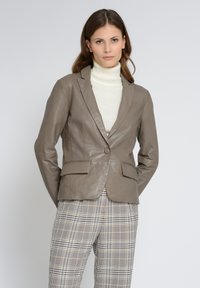 Läderblazer i taupe med enstaka knappstängning, slagkrage och två framfickor, tillsammans med en krämfärgad polotröja och rutiga byxor.