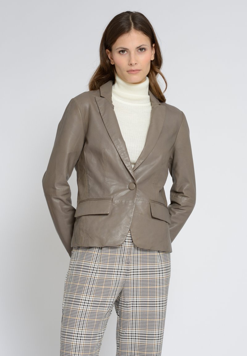 Läderblazer i taupe med enstaka knappstängning, slagkrage och två framfickor, tillsammans med en krämfärgad polotröja och rutiga byxor.