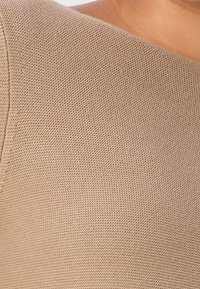 Gros plan sur un tissu beige texturé tricoté à l'épaule et au haut de la poitrine avec une couture visible et un fond de ton chair.
