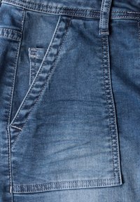 Tissu en denim de bleu moyen ; doté d'une poche et de coutures contrastantes. Texture lisse avec de légères variations de couleur.
