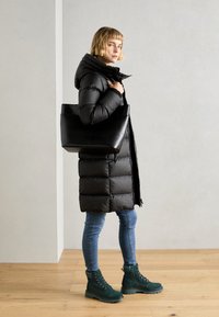 Manteau doudoune noir avec capuche, jean bleu et bottes vertes à lacets. Le modèle porte un sac fourre-tout structuré noir. Textures lisses et chaudes.