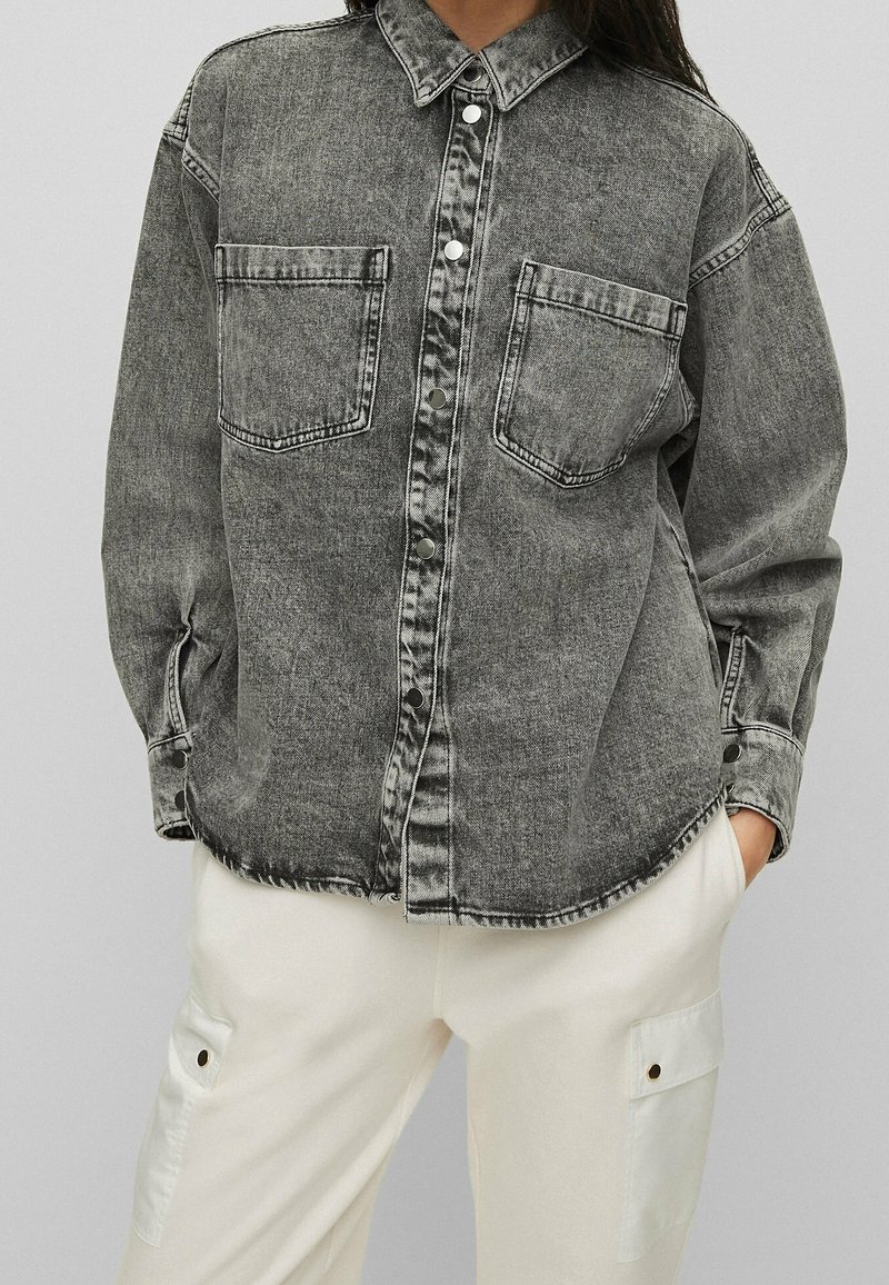 Chemise en denim gris avec deux poches avant, boutons pression, et surpiqûres blanches contrastantes, associée à un pantalon de couleur claire avec un détail de poche.