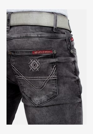 Jean en denim gris foncé avec une finition mate, ornés de coutures à étiquette rouge, de broderies uniques sur les poches et d'une ceinture beige contrastante.