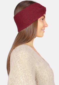 Bandeau tricoté bordeaux avec un design torsadé, fabriqué dans un tissu doux. Porté par un modèle aux longs cheveux bruns, mettant en valeur la texture.