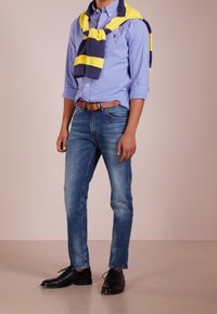 Camicia con bottoni a quadri blu, maglione a righe gialle e blu navy appoggiato sulle spalle, jeans denim blu e scarpe nere con lacci.