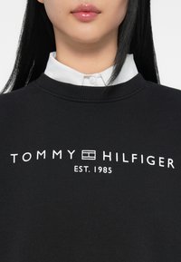 Juodas sportinis megztinis su apvaliu iškirpimu, kuriame balta spalva užrašyta "TOMMY HILFIGER" ir "EST. 1985" yra centriniu būdu išdėstyta priekyje, uždėta ant baltos marškinių apykaklės.
