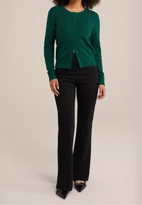 Grön, strukturerad kofta med rund halsringning, knäppning fram och långa ärmar, kombinerad med svarta, utsvängda byxor och spetsiga svarta skor.