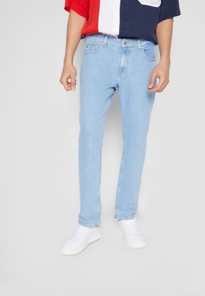 Mann trägt hellblaue Slim-Fit-Jeans, weiße Sneaker und ein kurzärmeliges Hemd mit roten, weißen und marineblauen Blockfarben, steht vor einem grauen Hintergrund.