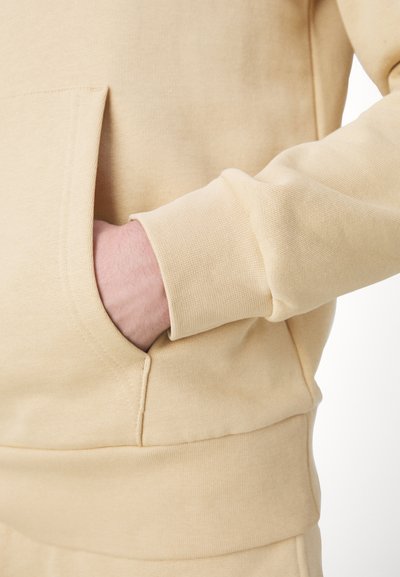 Sweat-shirt beige avec une poche kangourou, des poignets côtelés et une texture lisse. Présente une coupe décontractée et des détails cousus sur les coutures.