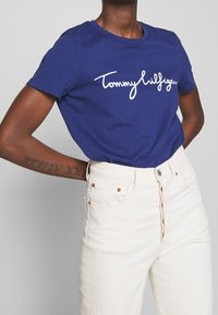 Námořnická modrá bavlněná trička s bílým logem "Tommy Hilfiger", krátké rukávy, kulatý výstřih, spárována s bílými džínami vysokého pasu.