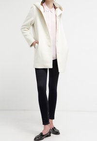 Manteau en laine blanche avec capuche, conçu avec une coupe droite et de gros boutons. Porté avec une chemise rose clair à col et des leggings noirs.