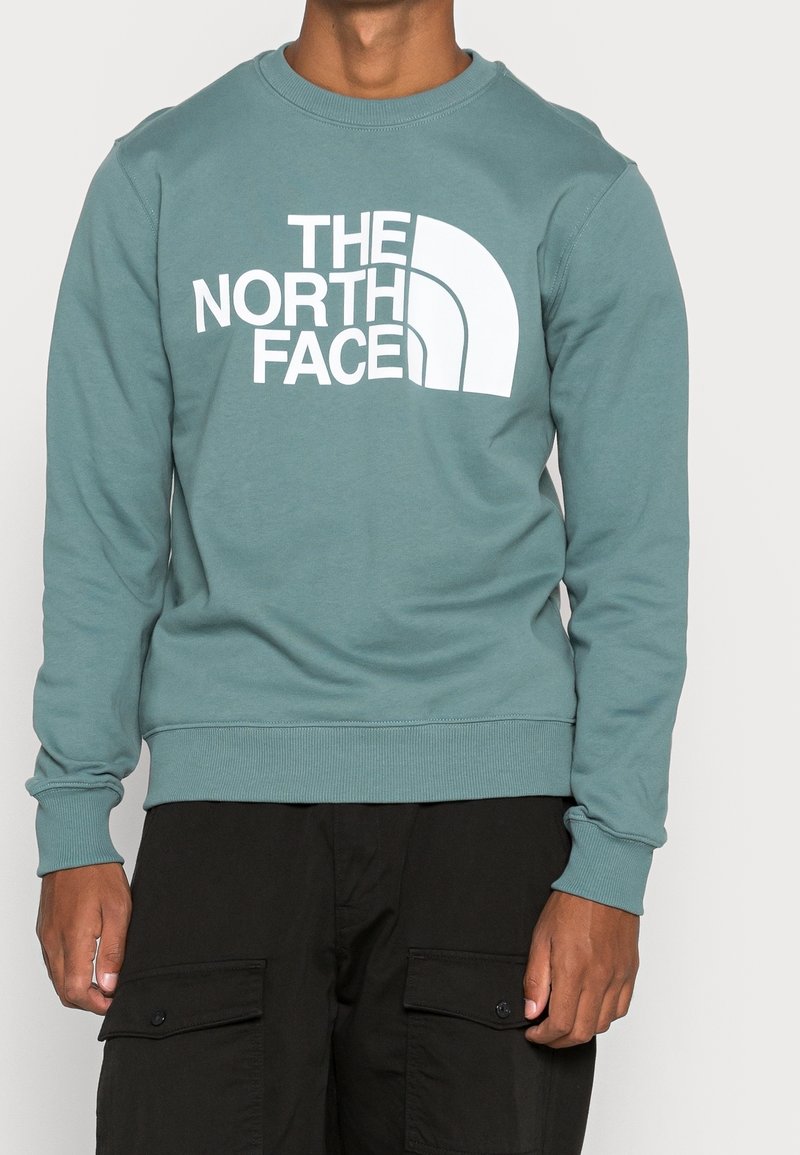 Man i en grönblå tröja med rund halsringning som har "The North Face"-logotypen i vitt och svarta cargo-byxor med knäppta fickor.