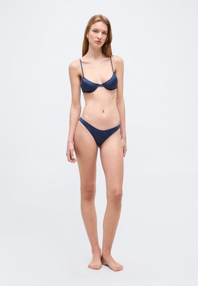 Νεαρή γυναίκα στέκεται ξυπόλυτη, φορώντας σκούρο μπλε μπικίνι, σε απλό ανοιχτό φόντο.