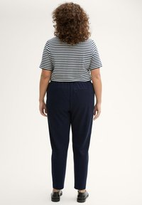 Mujer con cabello rizado que lleva una camiseta de rayas en blanco y negro de manga corta, pantalones azul marino y zapatos negros, de pie mirando hacia atrás.