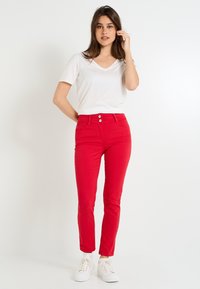 Jeans rojos de corte ajustado con cintura alta, cierre frontal de dos botones y blusa blanca de manga corta con escote en V, usados con zapatillas blancas.