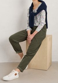 Hvid blomstret skjorte med lange ærmer, grønne bukser, en marineblå striktrøje bundet om halsen og hvide sneakers med brune accenter.