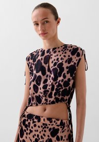 Tijgerprint top in licht taupe met donkere vlekken. Heeft een ronde halslijn, korte mouwen en verstelbare zijkanten voor een betere pasvorm. Glad textuur.