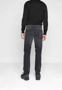 Man som står med ryggen mot kameran, klädd i svart långärmad skjorta, mörkgrå jeans med bakfickor och svarta sneakers framför en vit bakgrund.