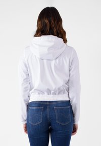 Giacca leggera bianca con cappuccio, vita elastica e maniche con polsini, abbinata a jeans blu con tasche posteriori e cuciture.