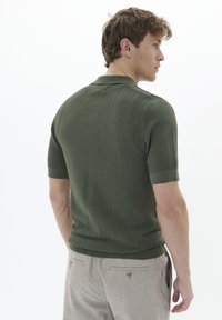 Polo cu mânecă scurtă în verde măsliniu, cu un model texturat în zigzag, guler și manșete ribbate, asortat cu pantaloni de culoare deschisă.