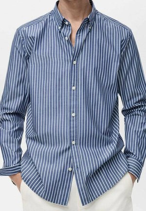 Man draagt een blauw overhemd met witte verticale strepen en een witte broek, handen in de zakken, tegen een effen achtergrond.