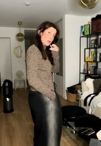 Vrouw met lang donker haar, draagend een bruine trui en een zwarte leren rok, staande in een woonkamer met boekenkasten en meubels.