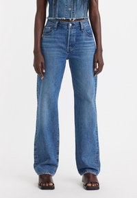Jeans de mezclilla azul de pierna ancha con una cintura alta, acabado deslavado, diseño clásico de cinco bolsillos y bordes sin dobladillo, combinados con sandalias negras.