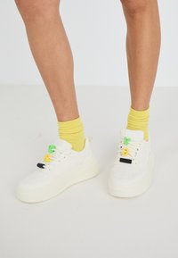 Weiße Sneakers mit einer kräftigen Sohle, verziert mit bunten Schnürsenkeln in Grün, Gelb und Schwarz. Getragen mit gelben gerippten Socken.