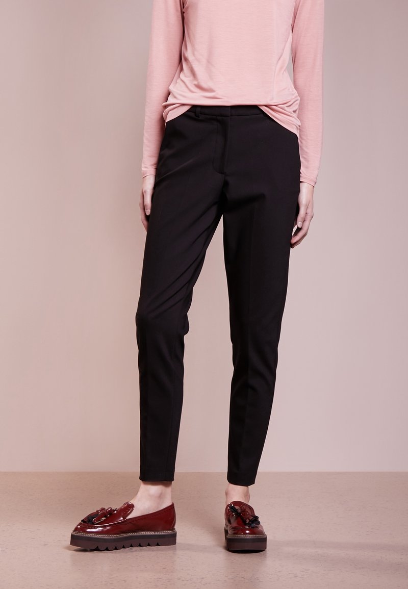 Pantalon noir sur mesure à coupe slim, associé à des mocassins compensés rouge foncé ornés d'un nœud décoratif et d'une semelle texturée. Haut à manches longues rose clair.