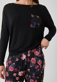Μαύρη μακριά μπλούζα με τσέπη με λουλουδάτο σχέδιο, συνδυασμένη με μαύρους παντελόνες που έχουν πολύχρωμο λουλουδάτο σχέδιο. Άνετη, χαλαρή εφαρμογή.