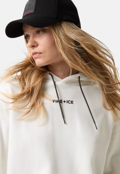 Młoda kobieta z długimi blond włosami, nosząca czarną czapkę i białą bluzę z kapturem z napisem "FIRE+ICE", patrząca w lewo na jednolitym tle.
