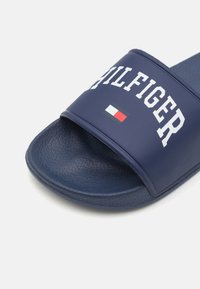 Chancleta de color azul marino hecha de goma texturizada. Presenta una tira ancha con "HILFIGER" en blanco y un pequeño acento de la bandera italiana.