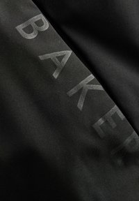 Čierna tkanina so slabo preplneným slovom "BAKER" v subtílnej, mierne priehľadnej tlači, ktorá vytvára elegantný, moderný vzhľad.