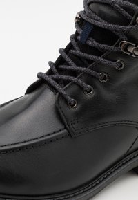 Stivaletto in pelle nera con lacci grigi, occhielli in metallo e cuciture dettagliate su uno sfondo bianco.