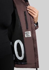 Paarse gewatteerde jas met zwarte voering, voorzien van een ritssluiting en een stoffen label met de tekst "Een warme omhelzing gemaakt van stof."