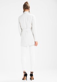 Biały płaszcz z paskiem, długie rękawy, widok od tyłu. Noszony z dopasowanymi białymi spodniami i czarnymi sandałami na wysokim obcasie. Gładka faktura.