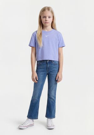 Mladé dievča s dlhými blond vlasmi, oblečené v svetlofialovom crop tope značky Nike, modré džínsy a biele tenisky, stojace na bielom pozadí.