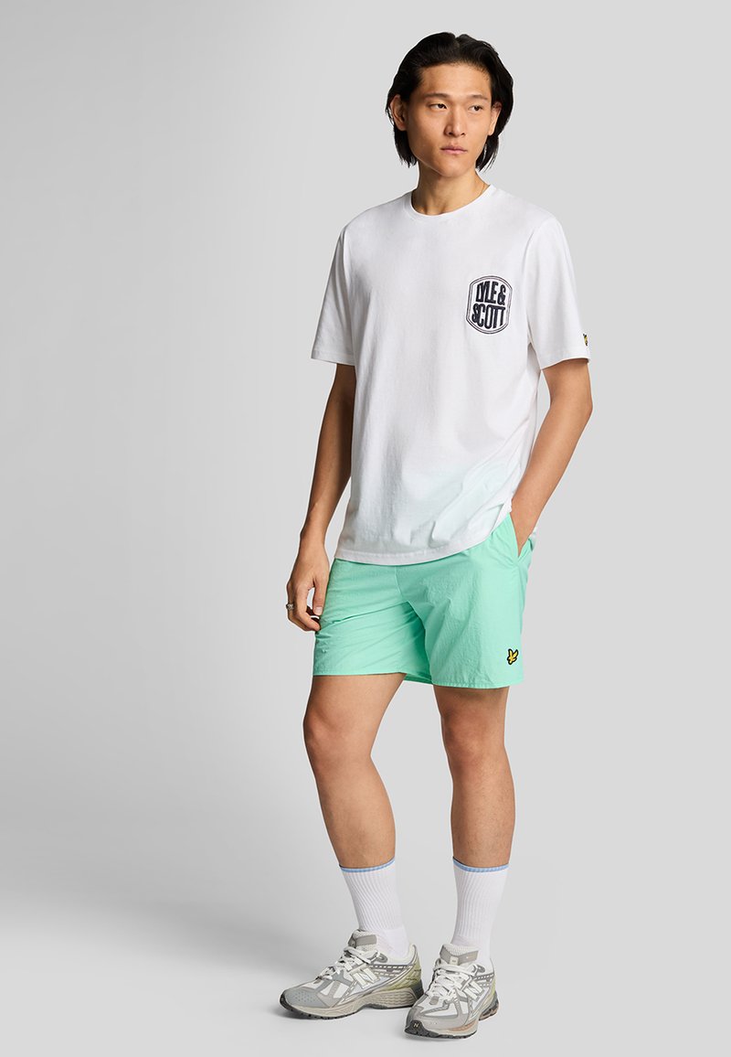 Maglietta bianca in cotone con logo nero sul petto, abbinata a pantaloncini verde menta con logo, calzini atletici bianchi e scarpe.