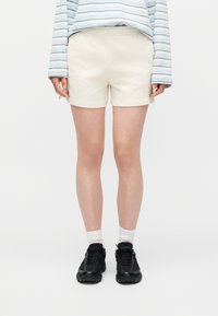 Άτομο που φοράει βερμούδα εκρού, μαύρα αθλητικά παπούτσια, λευκές κάλτσες και μακρυμάνικο μπλε και άσπρο ριγωτό πουκάμισο, στέκεται μπροστά σε λευκό φόντο.