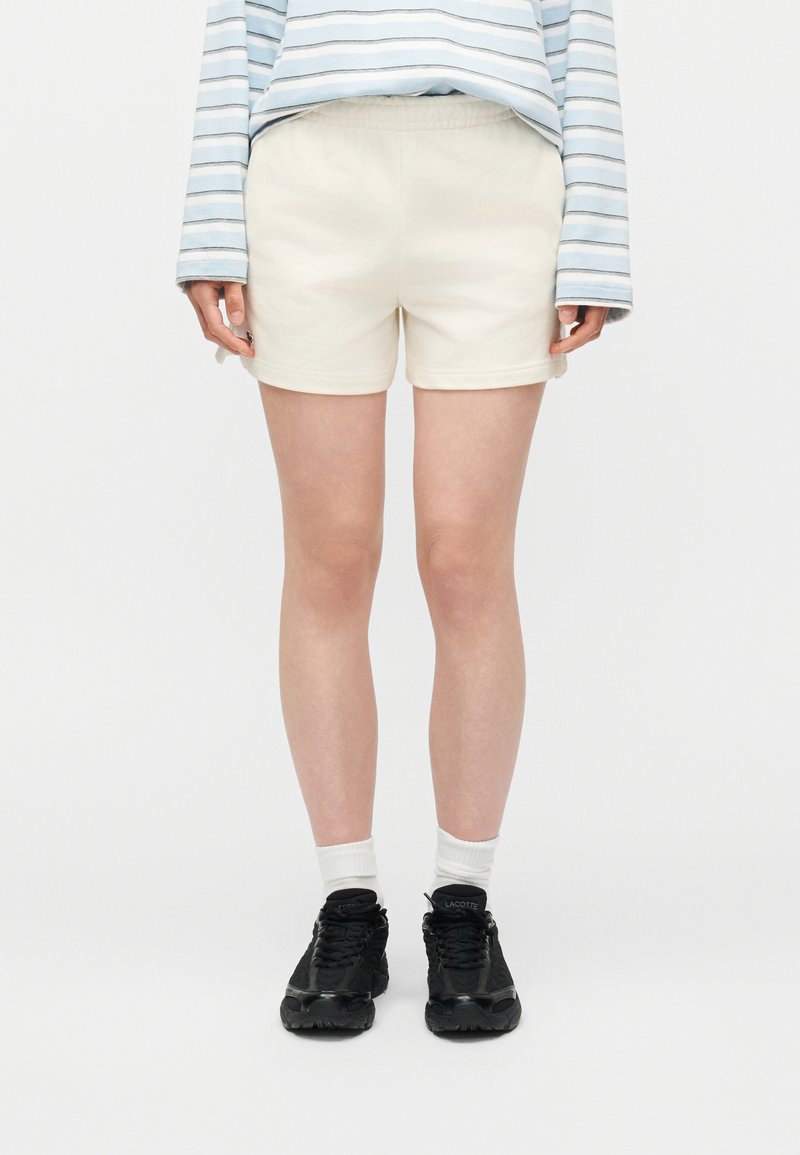 Άτομο που φοράει βερμούδα εκρού, μαύρα αθλητικά παπούτσια, λευκές κάλτσες και μακρυμάνικο μπλε και άσπρο ριγωτό πουκάμισο, στέκεται μπροστά σε λευκό φόντο.