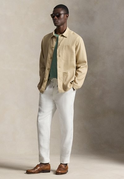 Άνδρας με σκούρα γυαλιά ηλίου, μπεζ σακάκι με κουμπιά, πράσινο πουκάμισο, λευκό παντελόνι με κορδόνι και καφέ δερμάτινα μοκασίνια, στέκεται μπροστά σε ουδέτερο φόντο.