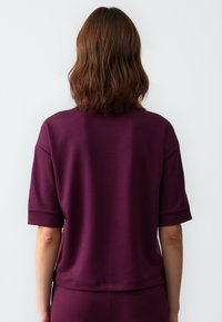 Korte mouwen, ronde hals top van diep bordeaux stof. Heeft een losse pasvorm met omgeslagen manchetten en een iets cropped zoom. Glad textuur.