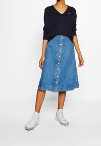 Femme portant un pull à col en V bleu marine et une jupe midi en denim bleu avec des baskets montantes blanches.
