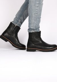 Stivali alla caviglia in pelle nera con finitura testurizzata, zip laterale e suola in gomma. Accento marrone attorno alla suola. Indossati con jeans azzurri.