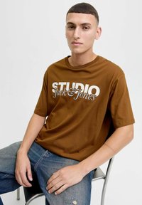 Brązowy bawełniany t-shirt z wyrazistym białym napisem "STUDIO Jack & Jones". Model siedzi, nosząc zniszczone niebieskie dżinsy z widocznymi detalami na rękach.