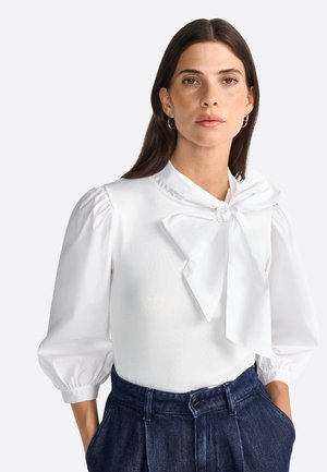 Weiße Bluse mit Puffärmeln und einer großen Schleife am Kragen, strukturierter Stoff, kombiniert mit dunkelblauen, hoch taillierten Hosen.