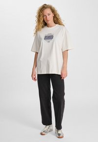 Crème korte mouw t-shirt met grafische print op de voorkant, gecombineerd met zwarte wijde jeans en zilveren sportschoenen met tan kleurige zolen.