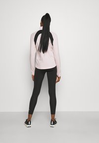 Langärmliges, hellrosa Sportoberteil mit schwarzen Schulterakzenten, kombiniert mit schwarzen Leggings und schwarzen Sneakers, von hinten gesehen.